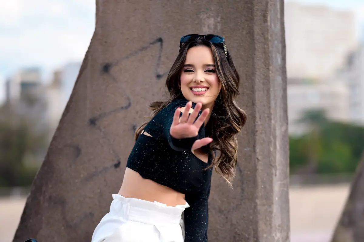 Mulher sorrindo e acenando, sentada num muro com a cidade ao fundo