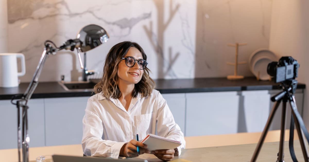 Mulher escrevendo num bloco de notas à mesa, com notebook e câmera ao lado